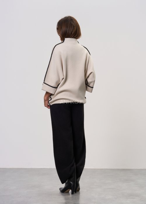 Beżowa damska bluzka o luźnym kroju, z półgolfem i rękawami 3/4, wykończona kontrastową czarną lamówką wzdłuż ramion, rękawów i boków oraz ozdobnymi guzikami przy mankietach.