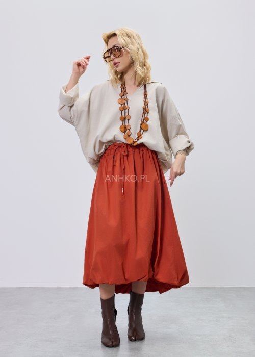 Beżowa bluzka damska oversize z dekoltem V i podwijanymi rękawami, zestawiona z ceglastą spódnicą w casualowej stylizacji.