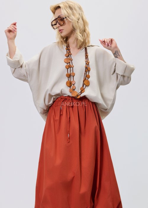 Beżowa bluzka damska oversize z dekoltem V i podwijanymi rękawami, zestawiona z ceglastą spódnicą w casualowej stylizacji.