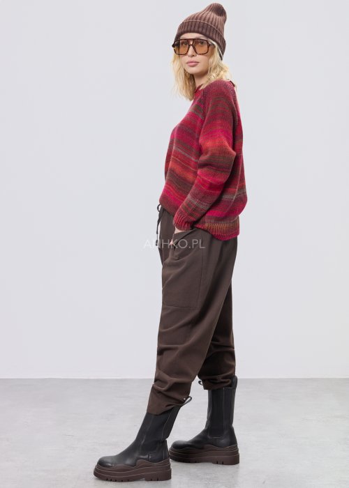 Bordowy sweter damski w wielobarwne paski w odcieniach ziemi, zestawiony z brązowymi spodniami w stylizacji casual na chłodniejsze dni.