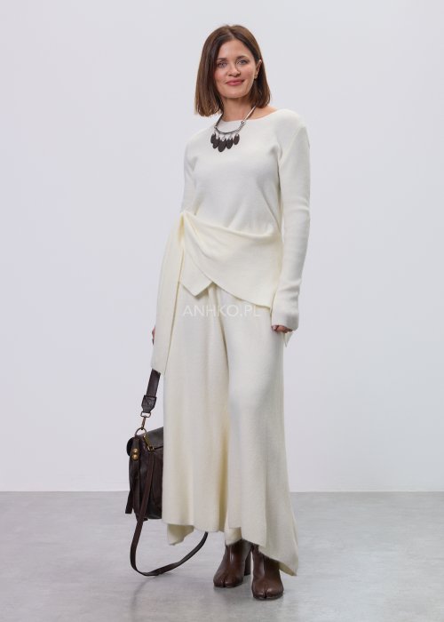 Śmietankowy komplet dzianinowy damski z asymetryczną tuniką i szerokimi spodniami, stylizowany z brązowymi botkami i torebką w minimalistycznym wnętrzu.