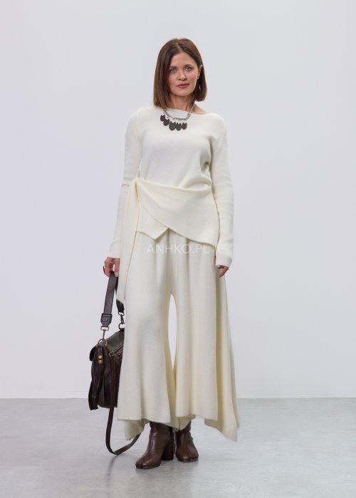 Śmietankowy komplet dzianinowy damski z asymetryczną tuniką i szerokimi spodniami, stylizowany z brązowymi botkami i torebką w minimalistycznym wnętrzu.