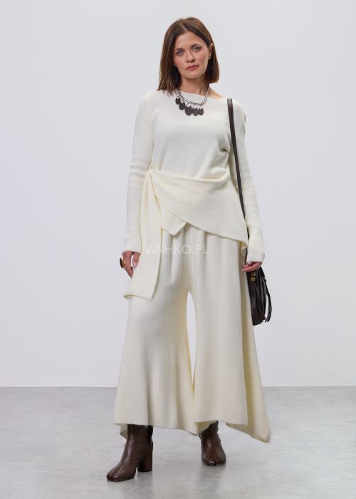 Śmietankowy komplet dzianinowy damski z asymetryczną tuniką i szerokimi spodniami, stylizowany z brązowymi botkami i torebką w minimalistycznym wnętrzu.