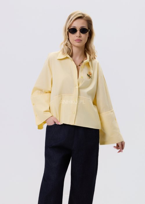 Jasnożółta koszula damska Velin Soft o luźnym kroju z klasycznym kołnierzykiem, dłuższym tyłem i poszerzanymi rękawami, stylizowana z ciemnymi spodniami.