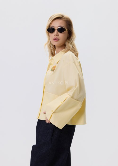 Jasnożółta koszula damska Velin Soft o luźnym kroju z klasycznym kołnierzykiem, dłuższym tyłem i poszerzanymi rękawami, stylizowana z ciemnymi spodniami.