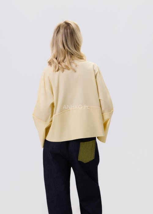 Jasnożółta koszula damska Velin Soft o luźnym kroju z klasycznym kołnierzykiem, dłuższym tyłem i poszerzanymi rękawami, stylizowana z ciemnymi spodniami.