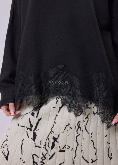 Czarna bluzka damska o luźnym kroju z dekoltem V i koronkowym wykończeniem u dołu, zestawiona z jasną spódnicą w casualowej stylizacji.