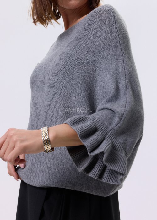 Szary sweter damski oversize z dekoracyjną falbaną na rękawie, gładka dzianina i luźny fason, stylizacja z czarnymi spodniami.
