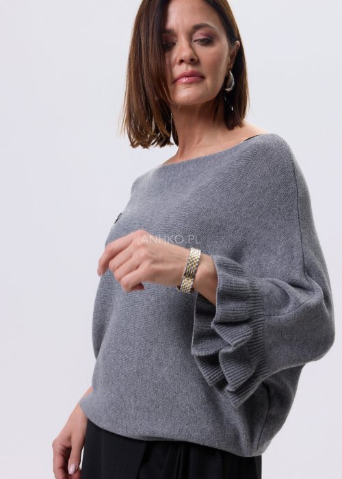 Szary sweter damski oversize z dekoracyjną falbaną na rękawie, gładka dzianina i luźny fason, stylizacja z czarnymi spodniami.