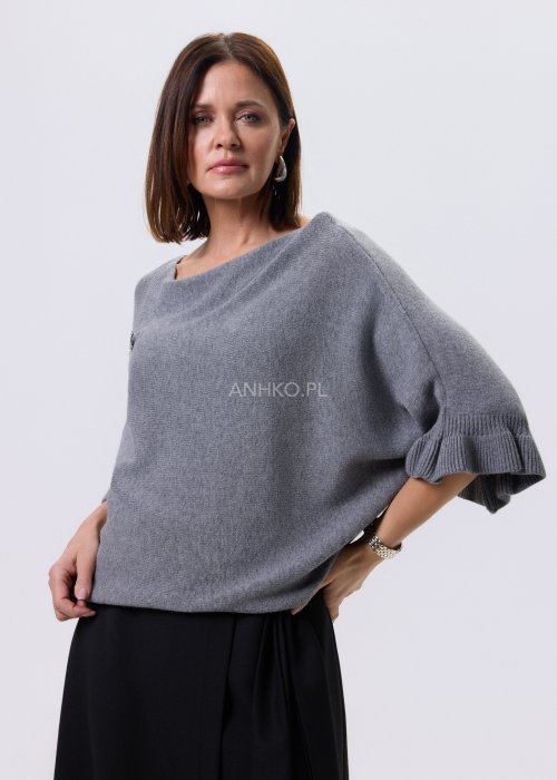 Szary sweter damski oversize z dekoracyjną falbaną na rękawie, gładka dzianina i luźny fason, stylizacja z czarnymi spodniami.