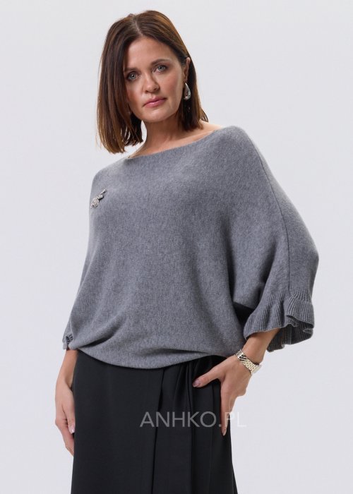 Szary sweter damski oversize z dekoracyjną falbaną na rękawie, gładka dzianina i luźny fason, stylizacja z czarnymi spodniami.
