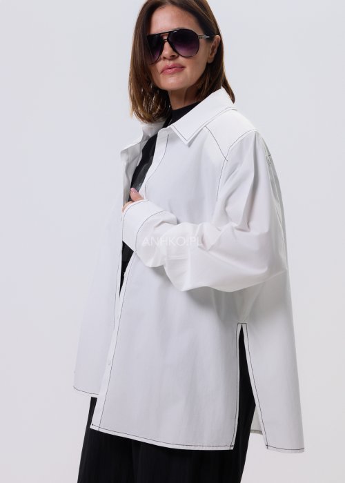 Biała koszula damska oversize z kontrastowym szwem i bocznymi rozcięciami, minimalistyczny fason, stylizacja z czarnymi spodniami.