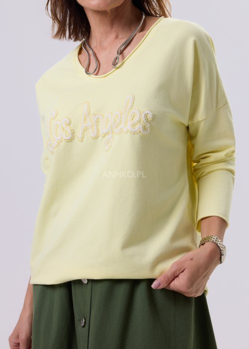 Żółta bluzka damska o luźnym kroju z haftem „Los Angeles” na przodzie, zestawiona z zieloną spódnicą w casualowej, miejskiej stylizacji.