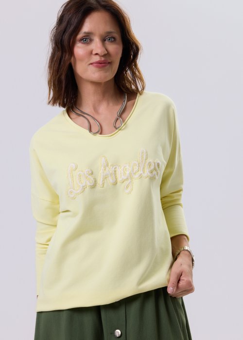 Żółta bluzka damska o luźnym kroju z haftem „Los Angeles” na przodzie, zestawiona z zieloną spódnicą w casualowej, miejskiej stylizacji.