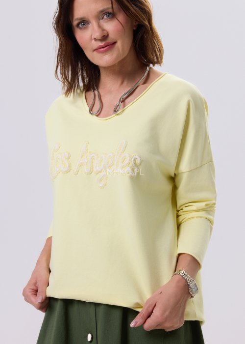 Żółta bluzka damska o luźnym kroju z haftem „Los Angeles” na przodzie, zestawiona z zieloną spódnicą w casualowej, miejskiej stylizacji.