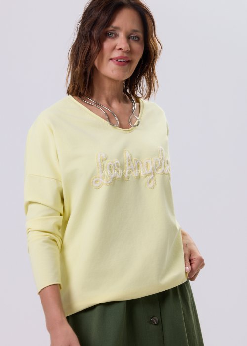 Żółta bluzka damska o luźnym kroju z haftem „Los Angeles” na przodzie, zestawiona z zieloną spódnicą w casualowej, miejskiej stylizacji.