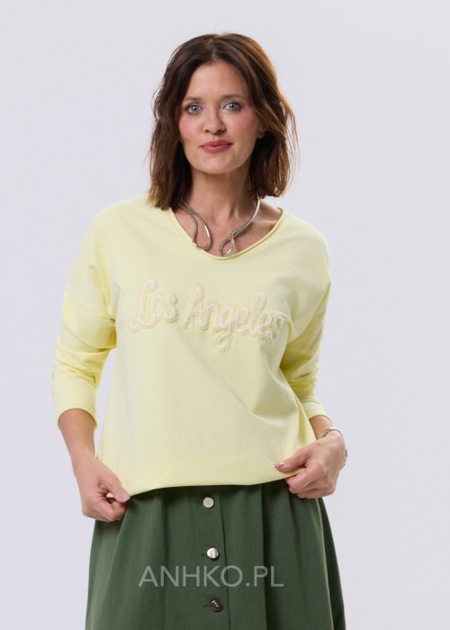 Żółta bluzka damska o luźnym kroju z haftem „Los Angeles” na przodzie, zestawiona z zieloną spódnicą w casualowej, miejskiej stylizacji.