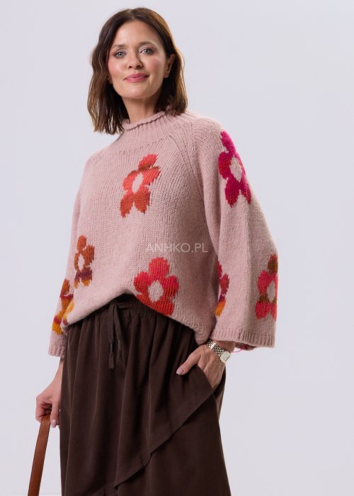 Pudrowo różowy sweter damski o luźnym kroju z dużymi, kolorowymi kwiatami w stylu retro, zestawiony z brązową spódnicą w casualowej stylizacji.