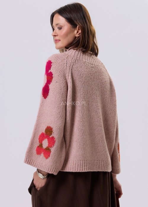 Pudrowo różowy sweter damski o luźnym kroju z dużymi, kolorowymi kwiatami w stylu retro, zestawiony z brązową spódnicą w casualowej stylizacji.