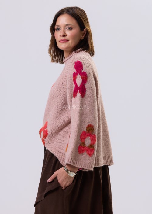 Pudrowo różowy sweter damski o luźnym kroju z dużymi, kolorowymi kwiatami w stylu retro, zestawiony z brązową spódnicą w casualowej stylizacji.