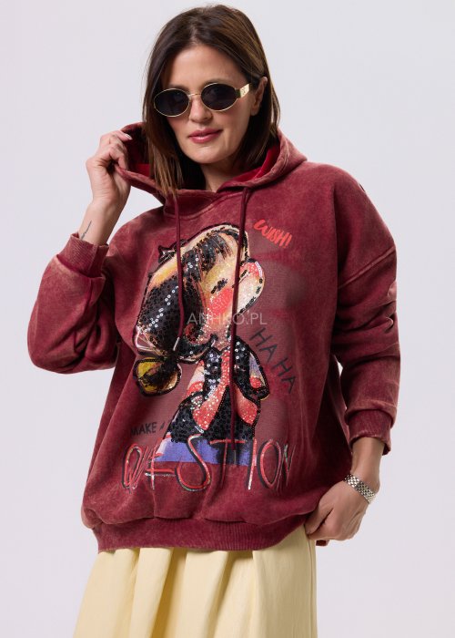 Bordowa bluza oversize z kapturem i dużą cekinową aplikacją na przodzie, zestawiona z jasną, długą spódnicą i czarnymi botkami w miejskiej stylizacji.