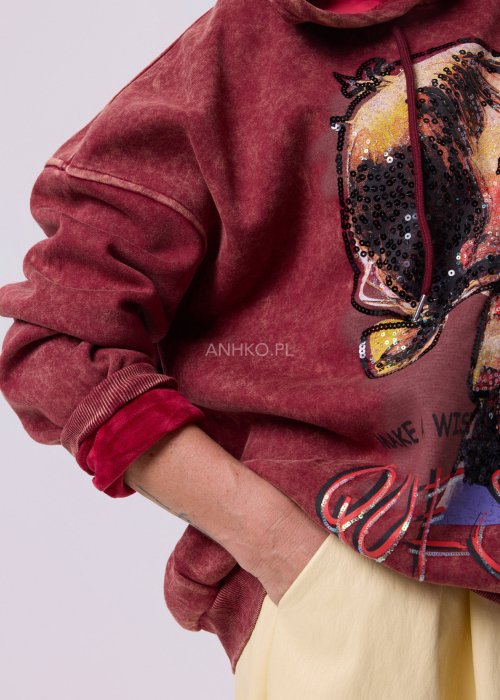 Bordowa bluza oversize z kapturem i dużą cekinową aplikacją na przodzie, zestawiona z jasną, długą spódnicą i czarnymi botkami w miejskiej stylizacji.
