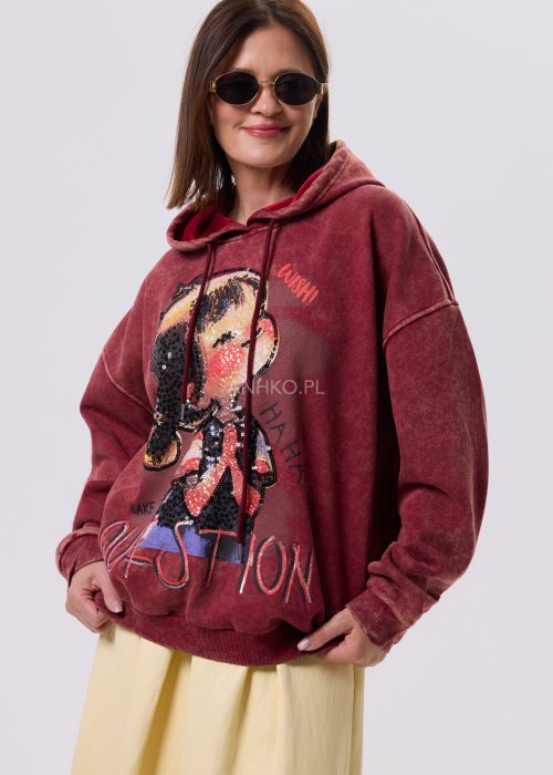 Bordowa bluza oversize z kapturem i dużą cekinową aplikacją na przodzie, zestawiona z jasną, długą spódnicą i czarnymi botkami w miejskiej stylizacji.