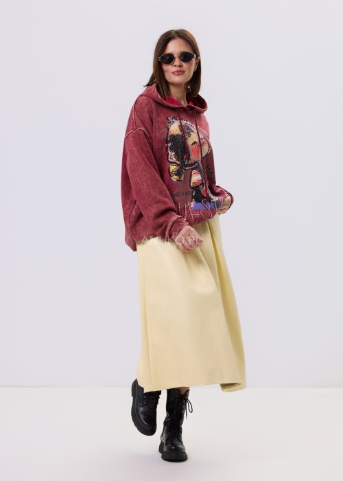 Bordowa bluza oversize z kapturem i dużą cekinową aplikacją na przodzie, zestawiona z jasną, długą spódnicą i czarnymi botkami w miejskiej stylizacji.