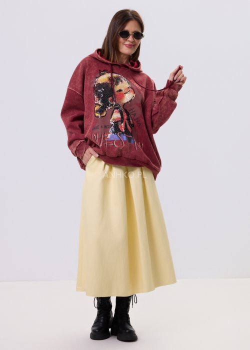 Bordowa bluza oversize z kapturem i dużą cekinową aplikacją na przodzie, zestawiona z jasną, długą spódnicą i czarnymi botkami w miejskiej stylizacji.