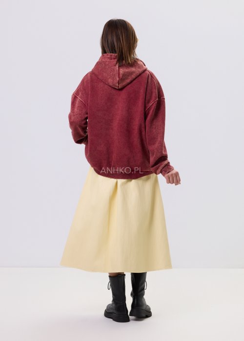 Bordowa bluza oversize z kapturem i dużą cekinową aplikacją na przodzie, zestawiona z jasną, długą spódnicą i czarnymi botkami w miejskiej stylizacji.
