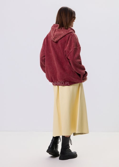 Bordowa bluza oversize z kapturem i dużą cekinową aplikacją na przodzie, zestawiona z jasną, długą spódnicą i czarnymi botkami w miejskiej stylizacji.