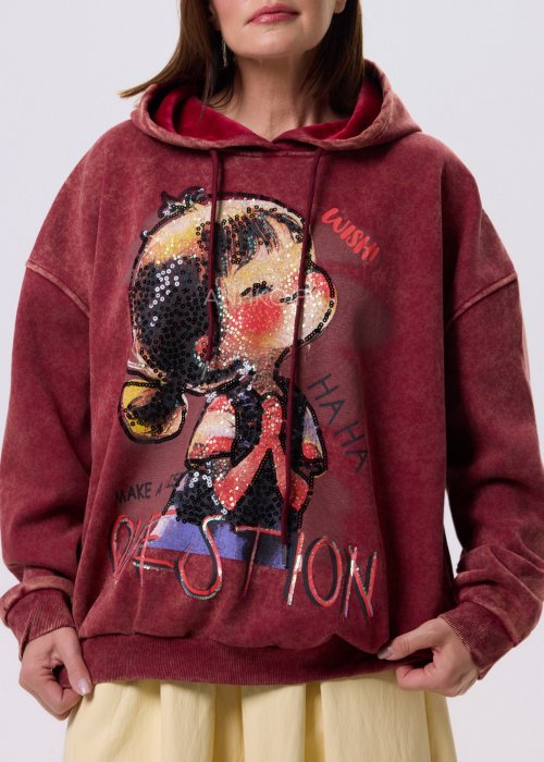 Bordowa bluza oversize z kapturem i dużą cekinową aplikacją na przodzie, zestawiona z jasną, długą spódnicą i czarnymi botkami w miejskiej stylizacji.