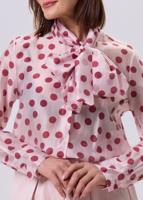 Jasnoróżowa bluzka Cherry Dot w wiśniowe groszki z wiązaną kokardą pod szyją, zapinana na guziki, lekki romantyczny fason.