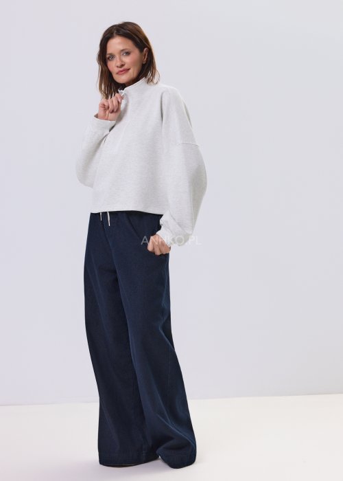 Jasnoszara damska bluza oversize z krótkim fasonem i stójką zapinaną na suwak, zestawiona z ciemnymi spodniami o szerokiej nogawce.