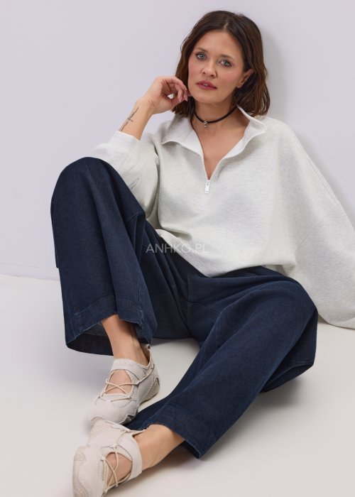 Jasnoszara damska bluza oversize z krótkim fasonem i stójką zapinaną na suwak, zestawiona z ciemnymi spodniami o szerokiej nogawce.