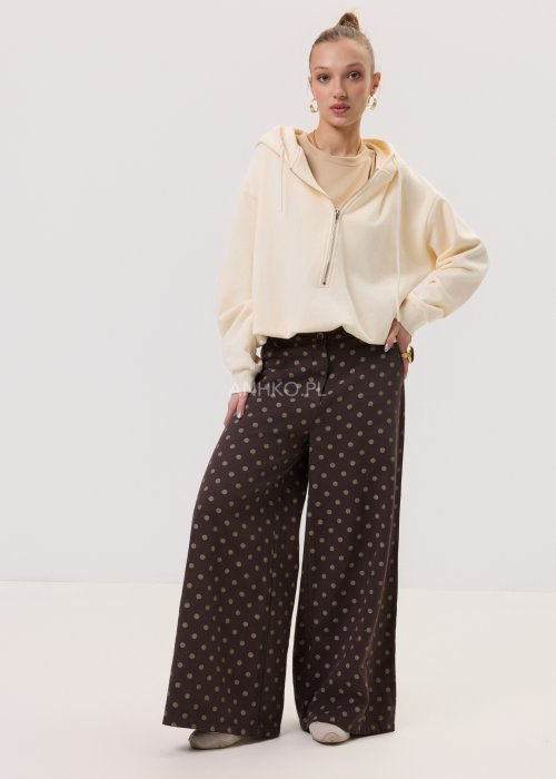 Brązowe damskie spodnie Retro Dots z szeroką nogawką, ozdobione beżowymi groszkami. Model z wysokim stanem i paskiem w talii, w stylizacji z kremowym topem oraz brązowymi botkami.