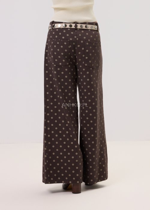 Brązowe damskie spodnie Retro Dots z szeroką nogawką, ozdobione beżowymi groszkami. Model z wysokim stanem i paskiem w talii, w stylizacji z kremowym topem oraz brązowymi botkami.