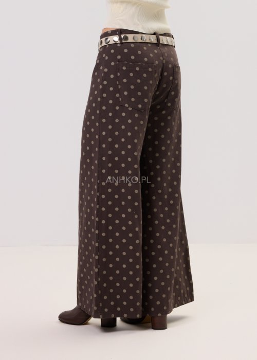 Brązowe damskie spodnie Retro Dots z szeroką nogawką, ozdobione beżowymi groszkami. Model z wysokim stanem i paskiem w talii, w stylizacji z kremowym topem oraz brązowymi botkami.