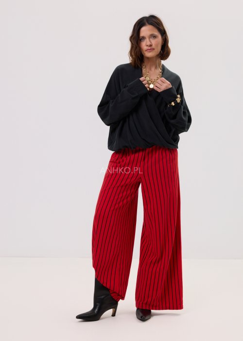 Czerwone spodnie damskie w czarne pionowe paski o szerokim kroju, zestawione z czarną bluzą i botkami na obcasie w nowoczesnej stylizacji.