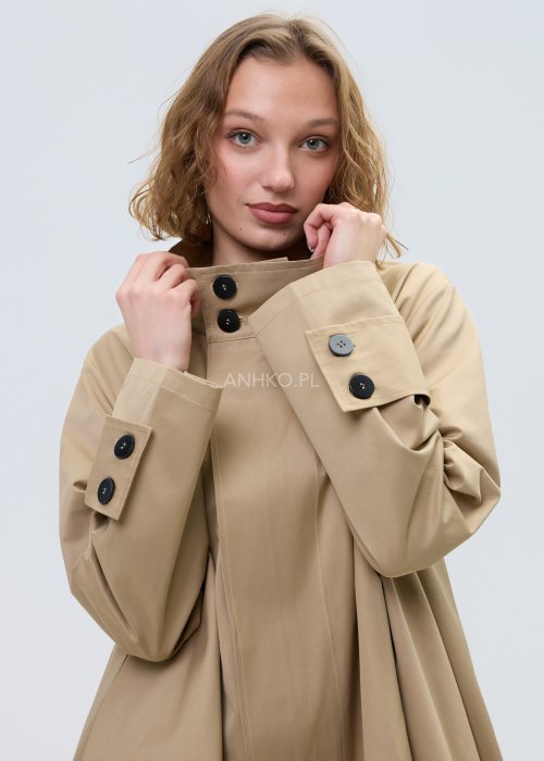 Beżowy płaszcz damski o luźnym, asymetrycznym kroju z wysokim kołnierzem. Model zapinany na zamek, z dekoracyjnymi guzikami przy kołnierzu i mankietach.