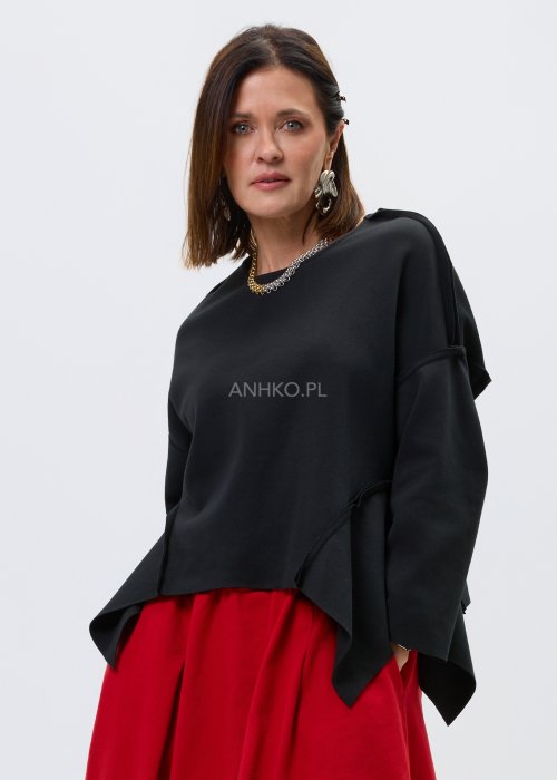 Czarna bluza oversize z szerokimi rękawami i asymetrycznym dołem, o luźnym, nowoczesnym kroju. Stylizacja z czerwoną spódnicą midi i botkami w wyrazistym, miejskim stylu.