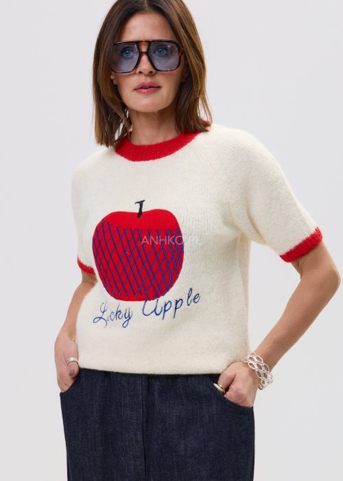 Kremowy sweter z czerwonymi mankietami i nadrukiem jabłka „Lucky Apple”, zestawiony z szerokimi jeansami i czerwonymi sandałami w modnej stylizacji.