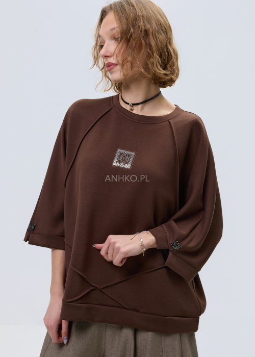 Brązowa damska bluza oversize z krótkim rękawem i zaokrąglonym dekoltem, model z ozdobną aplikacją na przodzie i dekoracyjnymi przeszyciami.