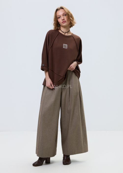 Brązowa damska bluza oversize z krótkim rękawem i zaokrąglonym dekoltem, model z ozdobną aplikacją na przodzie i dekoracyjnymi przeszyciami.