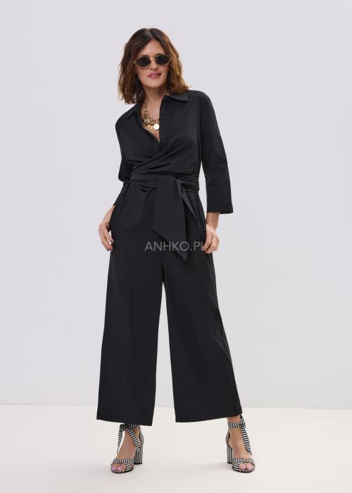 Czarny kombinezon damski z kopertową górą i paskiem w talii, z długim rękawem i szerokimi nogawkami, stylizowany z sandałami na obcasie.