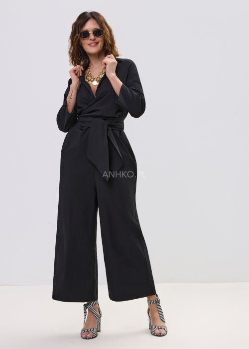 Czarny kombinezon damski z kopertową górą i paskiem w talii, z długim rękawem i szerokimi nogawkami, stylizowany z sandałami na obcasie.