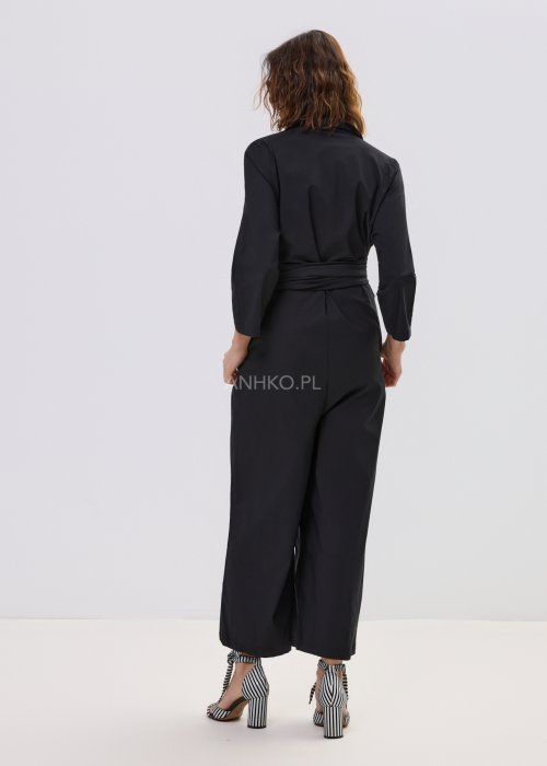 Czarny kombinezon damski z kopertową górą i paskiem w talii, z długim rękawem i szerokimi nogawkami, stylizowany z sandałami na obcasie.
