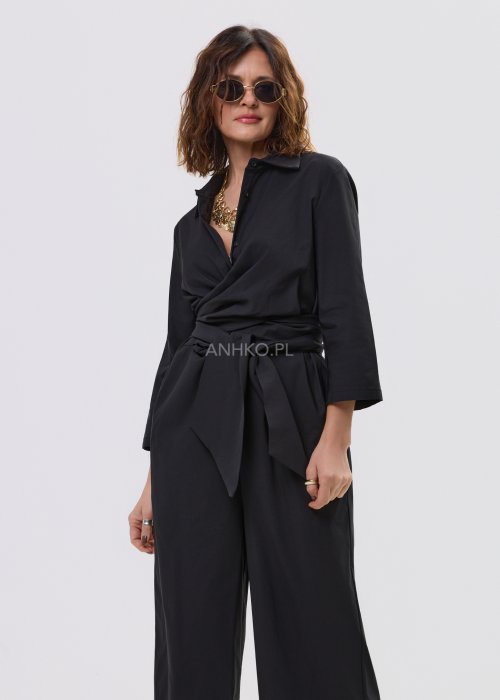 Czarny kombinezon damski z kopertową górą i paskiem w talii, z długim rękawem i szerokimi nogawkami, stylizowany z sandałami na obcasie.
