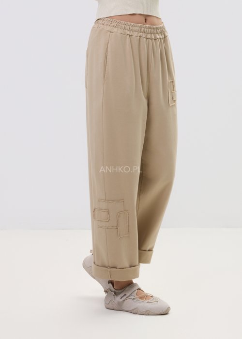 Beżowe spodnie damskie o luźnym kroju z elastycznym pasem i ozdobnymi naszyciami na nogawce.