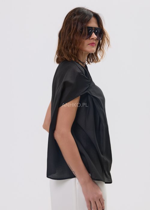 Czarna asymetryczna bluzka damska z lekkiej, półtransparentnej tkaniny, z efektownym drapowaniem na ramieniu i luźnym, swobodnie opadającym fasonem. Stylizacja z jasnymi spodniami.